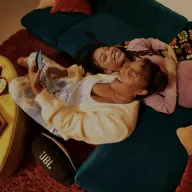 Two women of color relaxing on a teal couch, smiling while using a digital device with a JBL speaker visible on the floor, creating a cozy home entertainment scene.