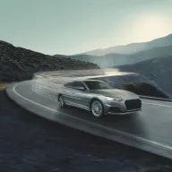 Bridgestone key visual with a sleek silver car speeds around a curvy mountain road, leaving light trails amid a serene mountainous backdrop