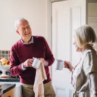 personalized-newsletters-v2.png - Image of a cheerful senior man in a burgundy sweater enjoying coffee in a kitchen while conversing with a woman, representing personalized retirement lifestyle content.