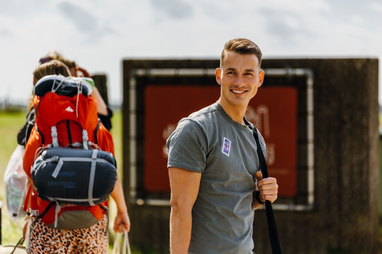 A smiling man in a gray T-shirt stands in the outdoors at Springbok's summer team event, holding a bag strap. Behind him, a woman carries a large backpack. 