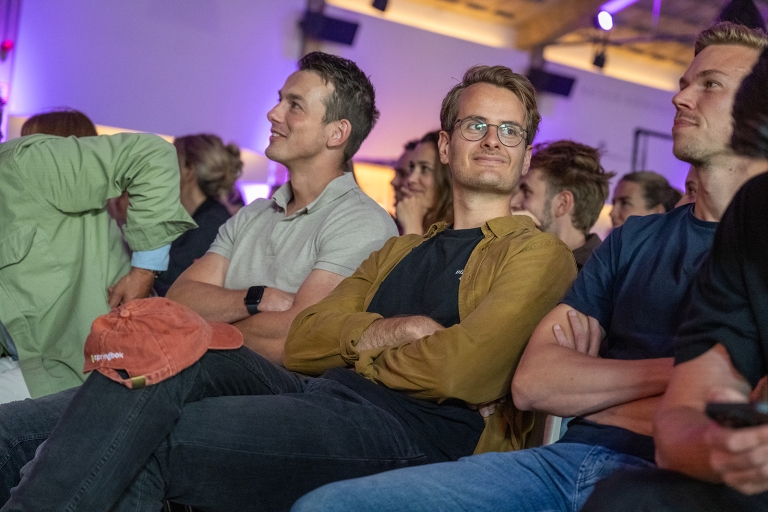 Audience members at Springbok's Dutch indoor team event sit closely in rows, attentively listening. The atmosphere is relaxed, with soft purple lighting.