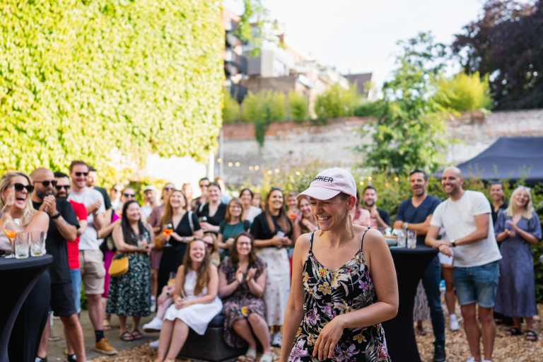A joyful woman in a floral dress and cap stands smiling amidst a clapping crowd at Springbok's summer barbecue event in Ghent, with a lush, green ivy wall and lights in the background.